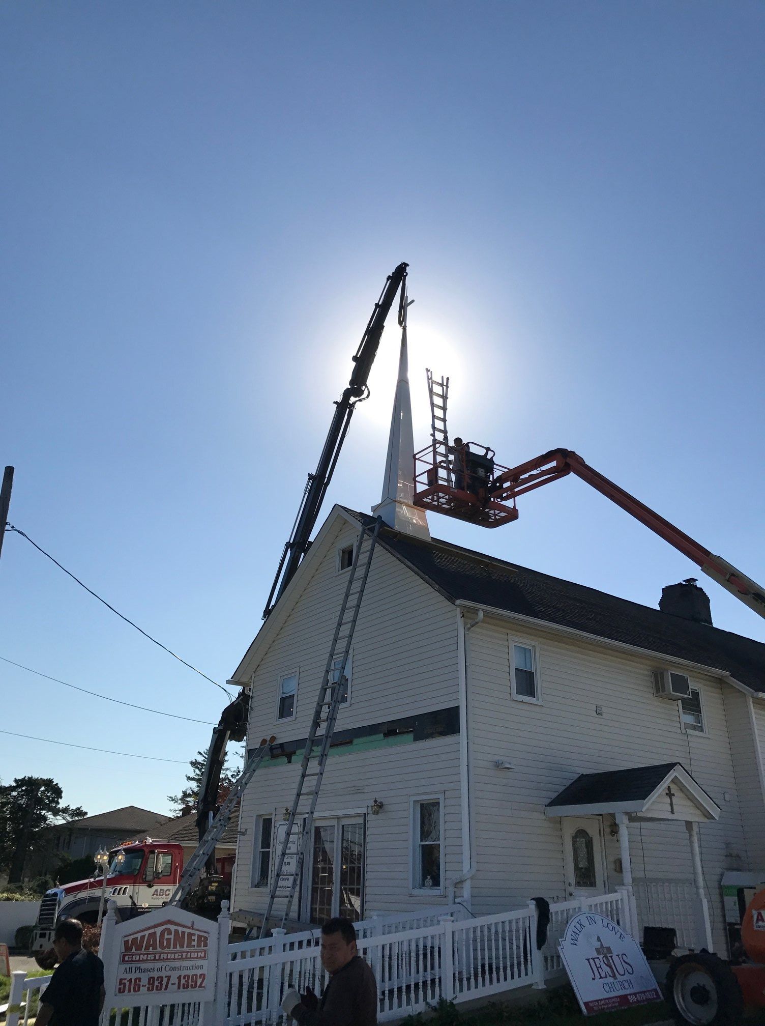 Church with steeple being worked on; two lift vehicles, bright sun.