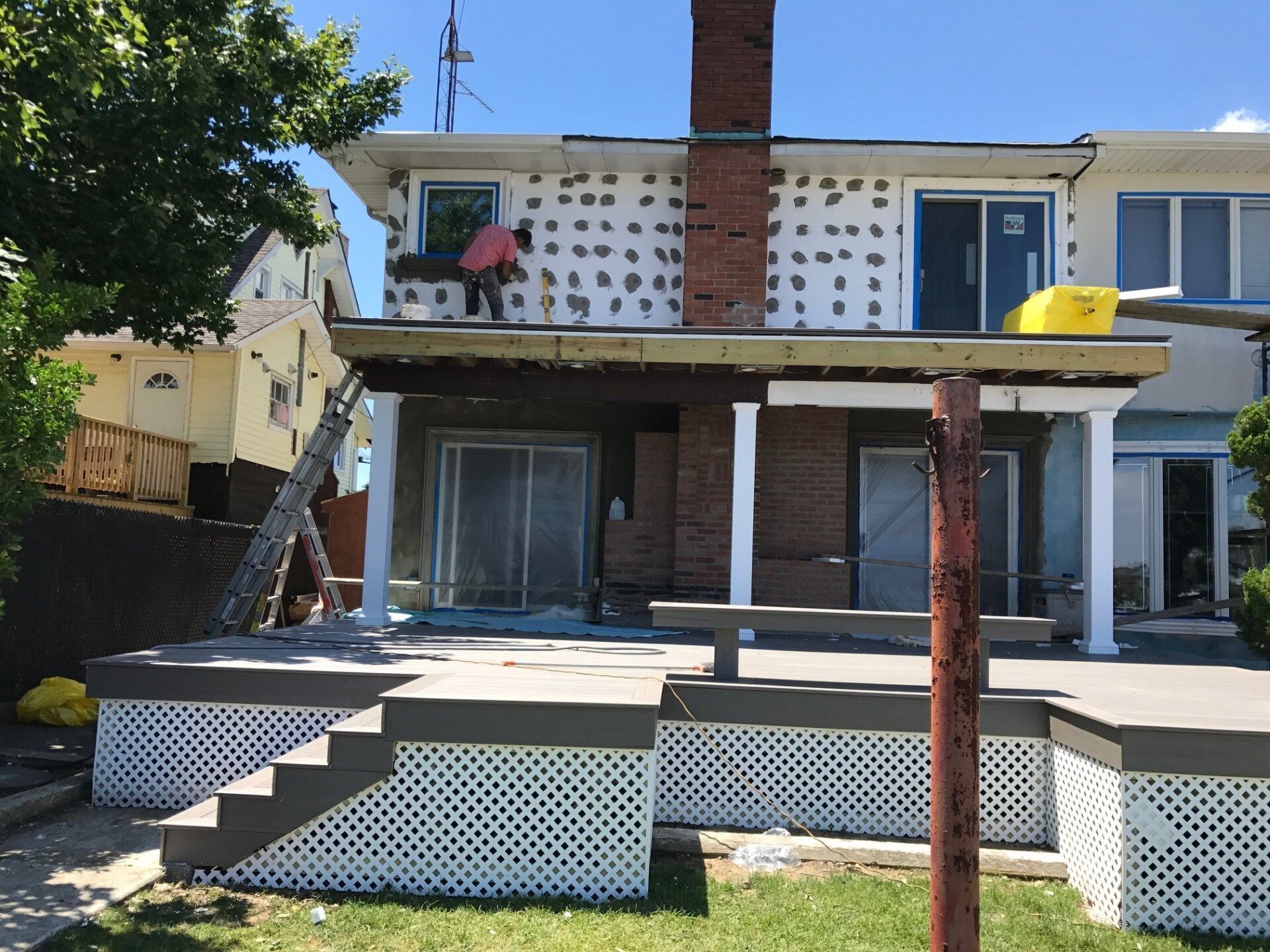 House exterior under renovation; worker on upper level, exposed siding. Brick chimney and porch, sunny day.