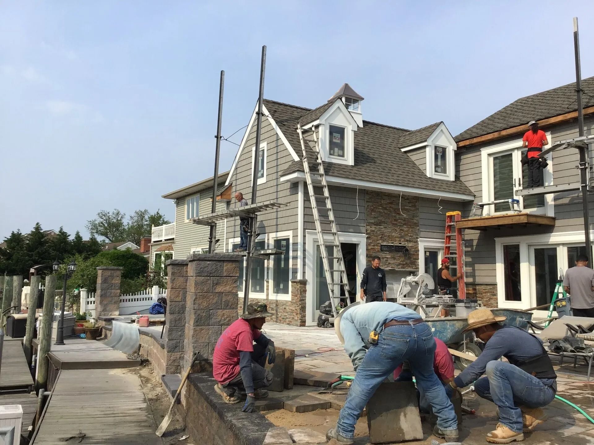 Construction workers installing stone on a house exterior near water. Scaffold and ladders are visible.