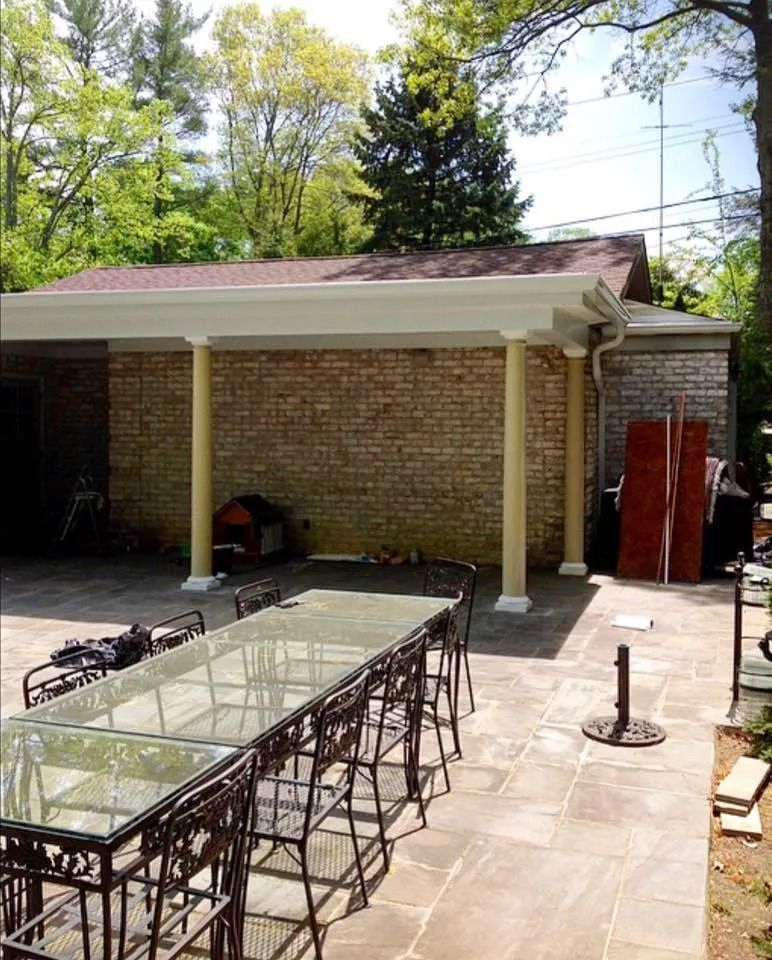Patio with long glass-top table and chairs under a covered area with brick wall and tan columns.
