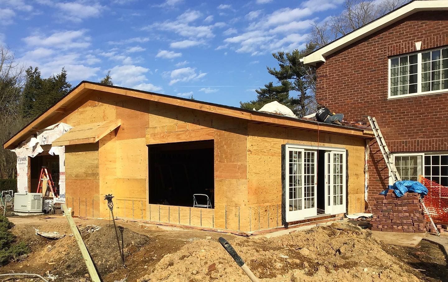 Garage addition under construction next to brick house. Raw wood framing, open door and window spaces, dirt.