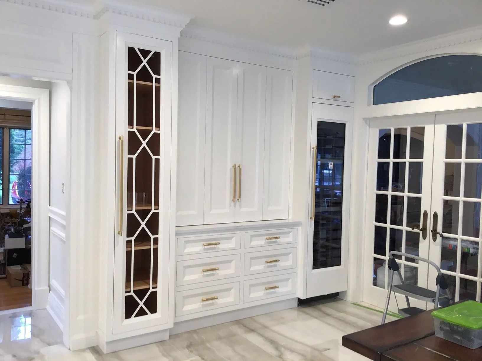 White kitchen cabinets with gold hardware and geometric glass-paneled display case, marble floor.
