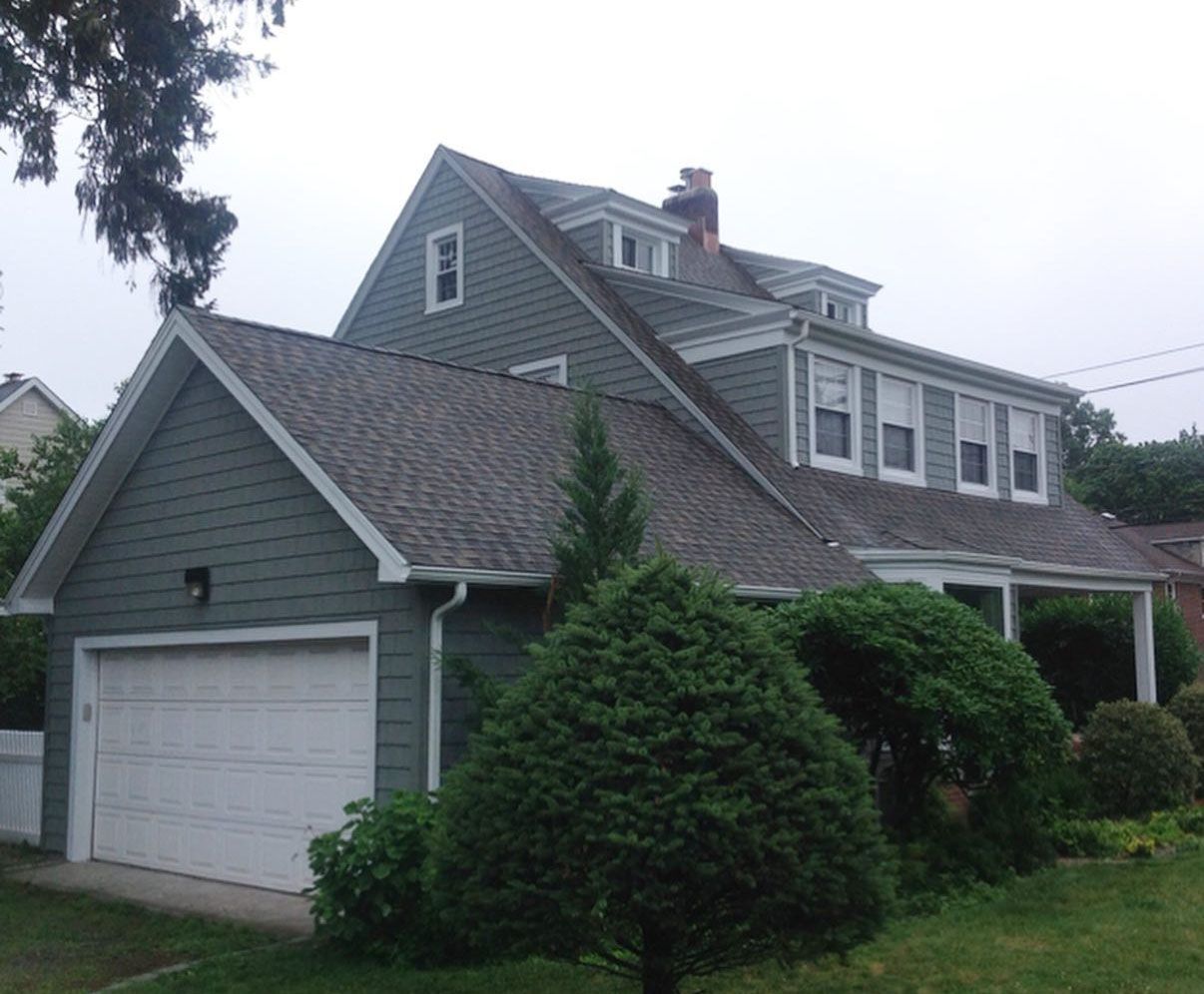 Green-sided house with a garage, gray roof, and white trim. Overcast day.