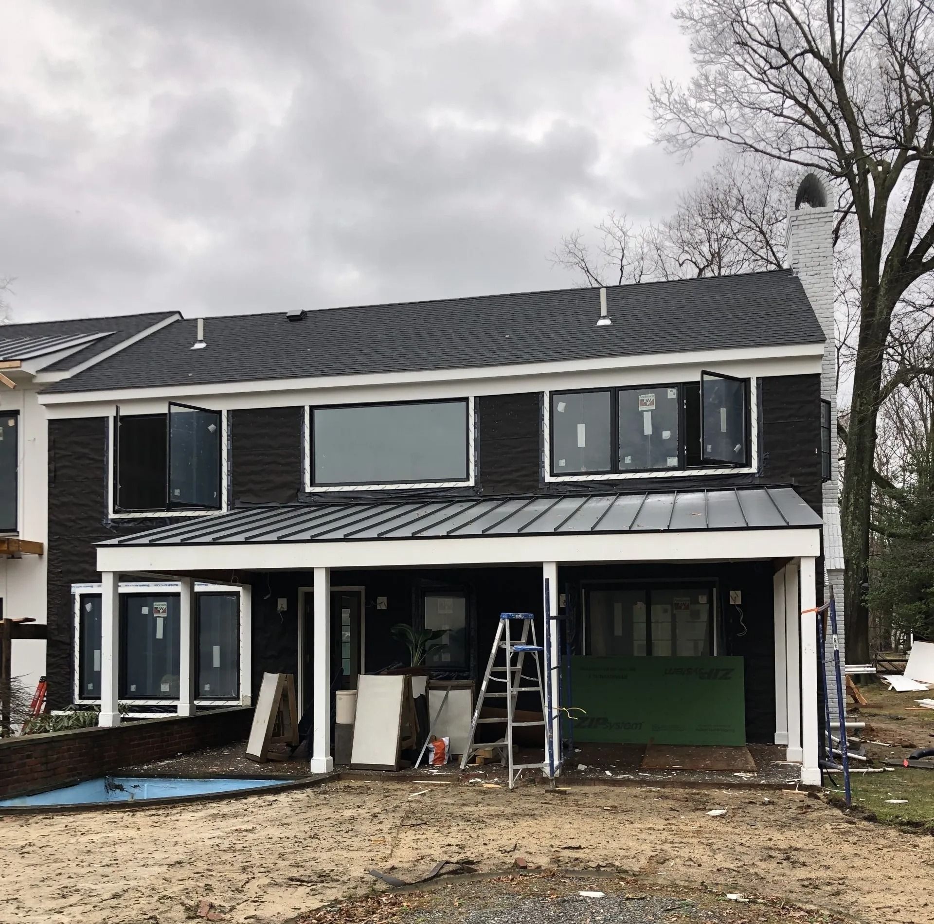 Black house under construction with large windows, covered patio, ladder, and a partially built pool.