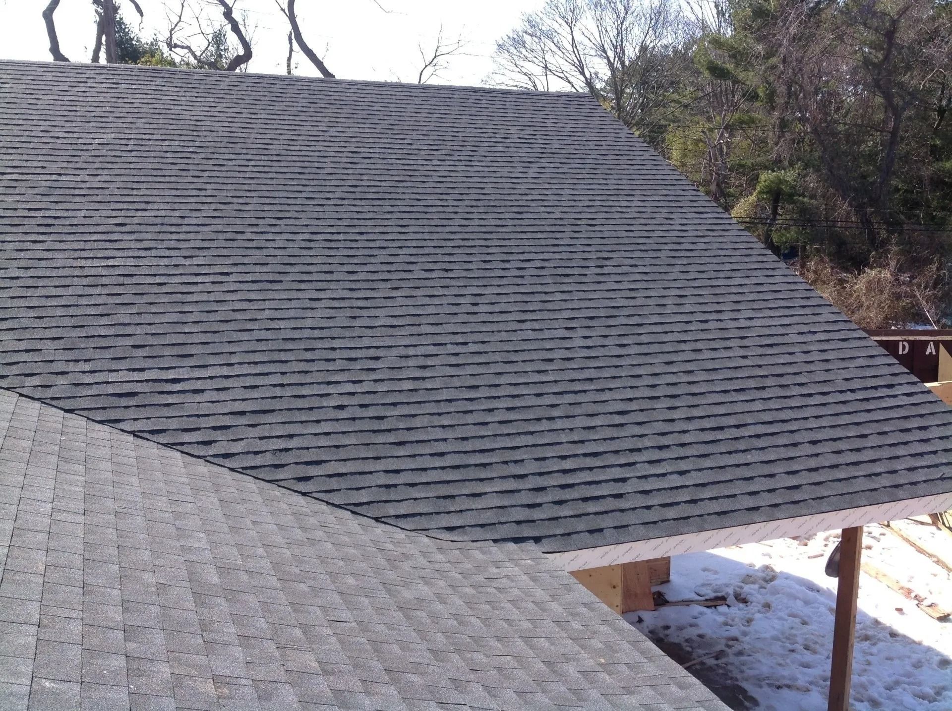 Gray asphalt shingle roof on a house, angled view. Sunny day, forest in background, some snow.