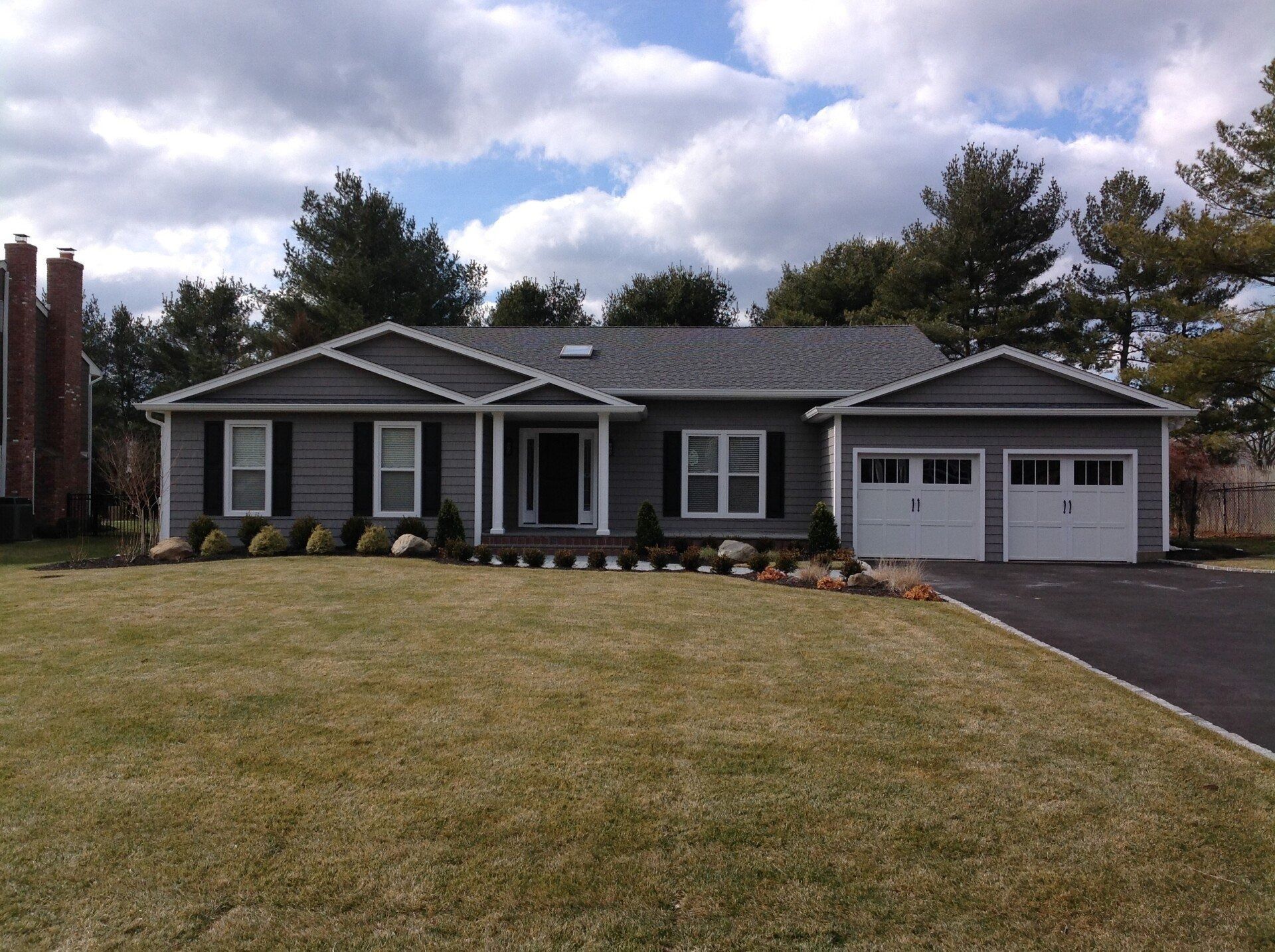 Gray ranch-style house with black shutters, white garage doors, and a green lawn.