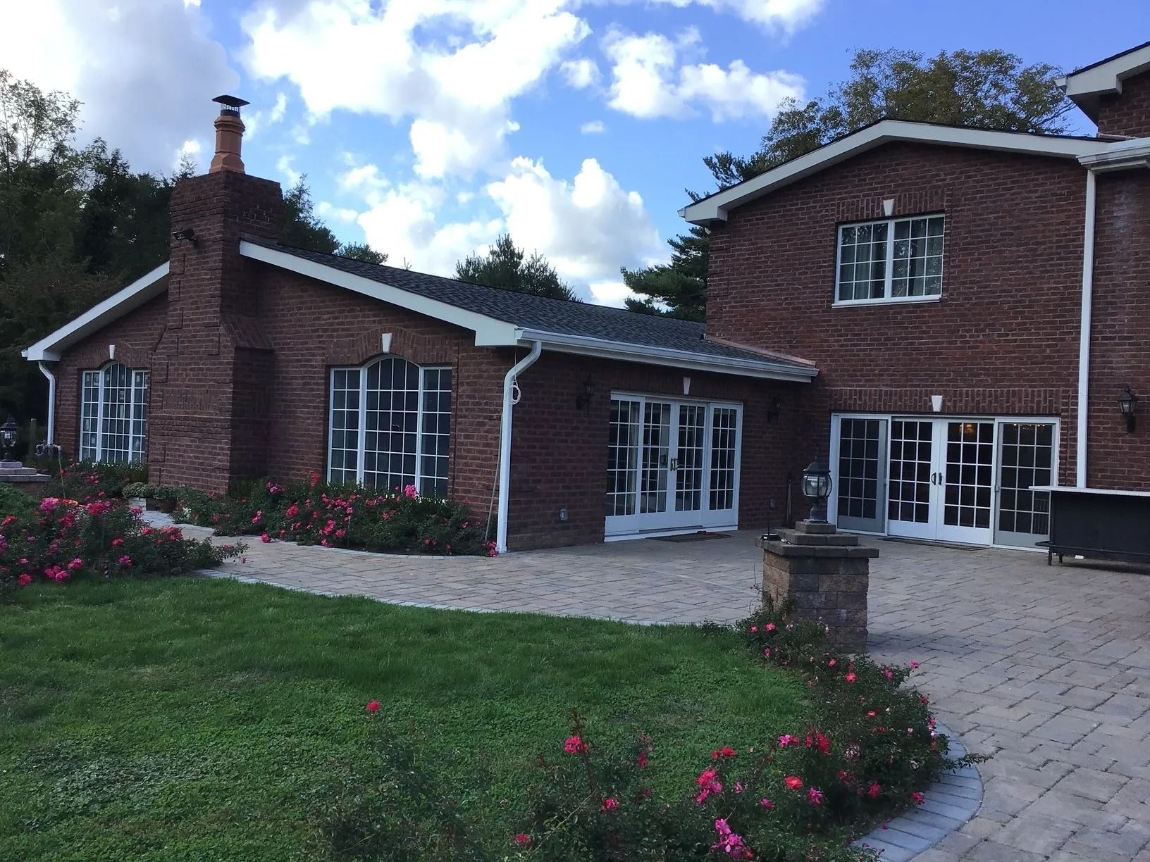Brick building with white-framed windows, surrounded by a paved patio and lawn with pink flowers, under a blue sky.