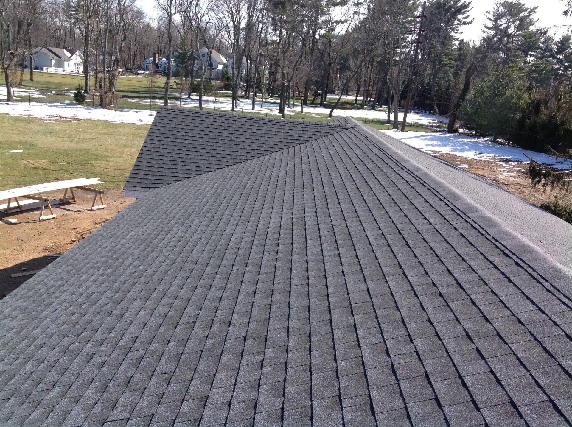Gray asphalt shingle roof on a house, angled view, with trees and snow in the background.