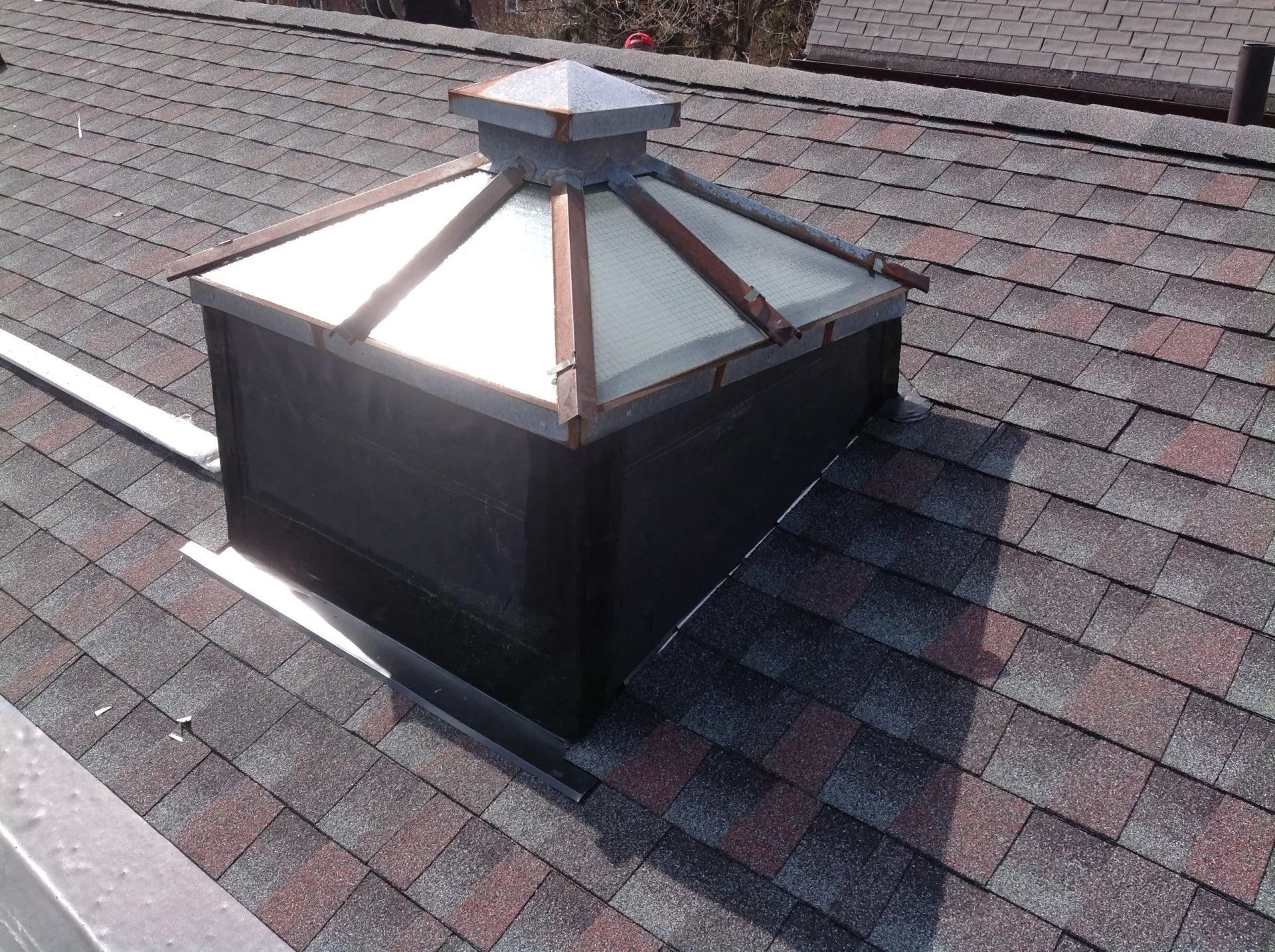 Skylight on a shingled roof, with a clear, peaked top and dark sides.