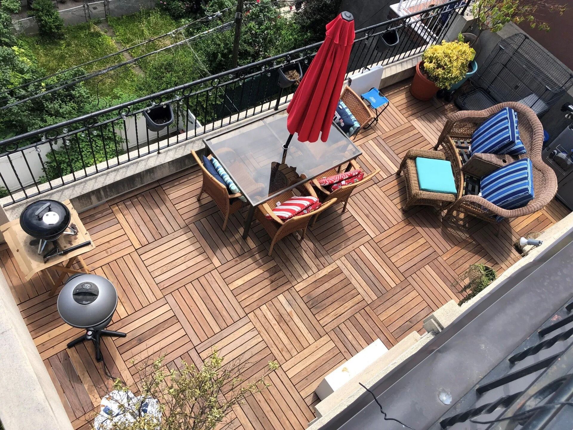 Outdoor deck with wood tiles, table with red umbrella, wicker seating, and a grill.
