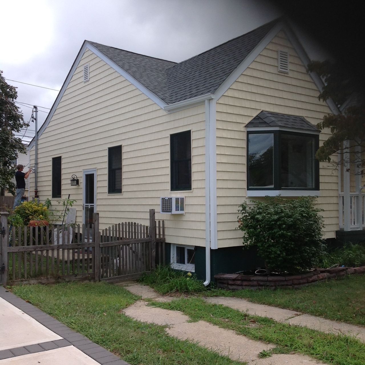 Yellow house with gray roof, black shutters, bay window, small fence, and driveway.