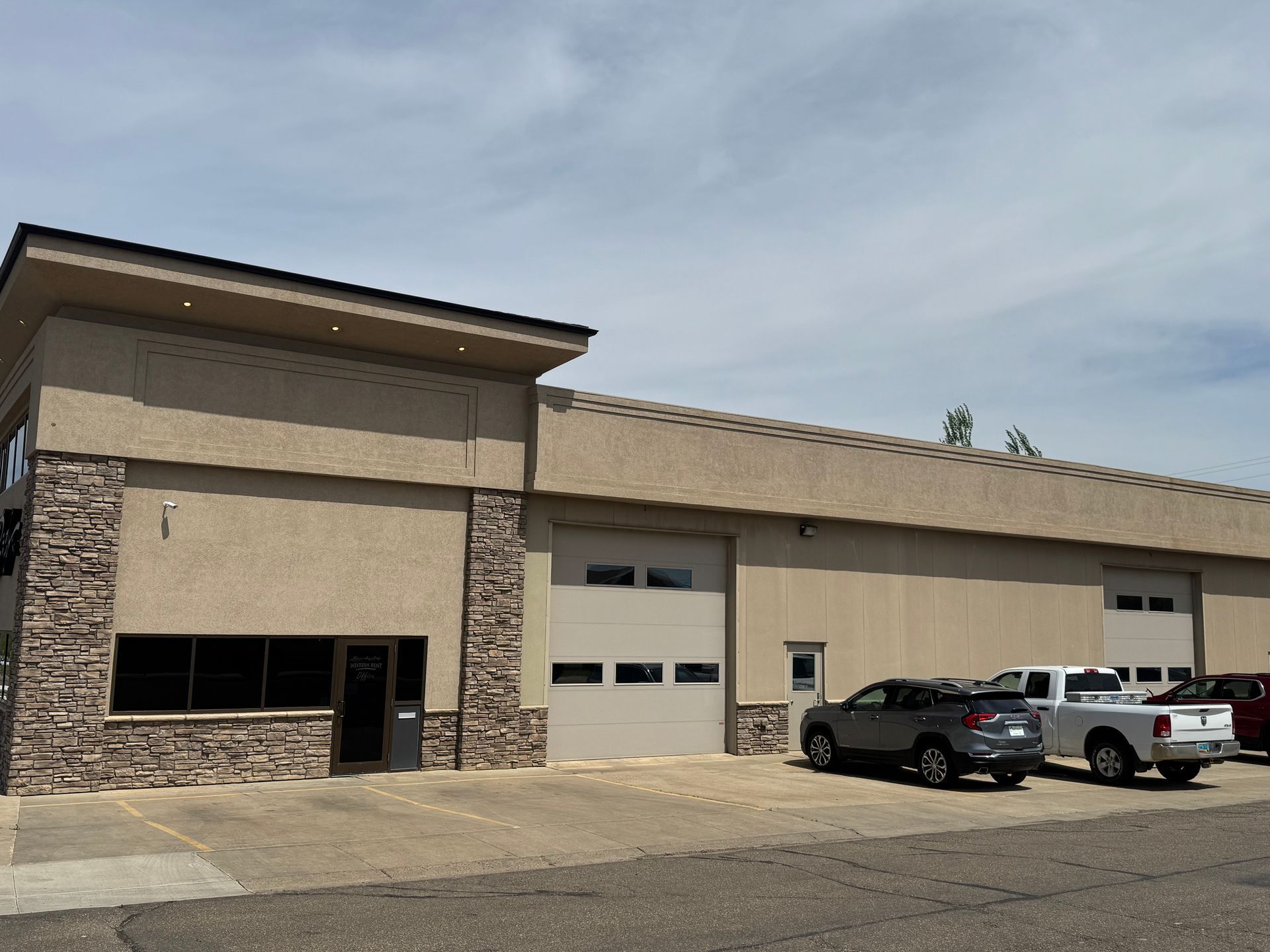Industrial building with beige walls, garage doors, and parked cars in front under a cloudy sky.