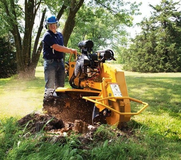 A person wearing safety gear uses a yellow stump grinder to remove a tree stump in a grassy, sunlit outdoor area.