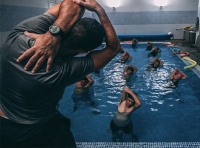 Swimming class in indoor pool, instructor demonstrating stretch while students follow in the water