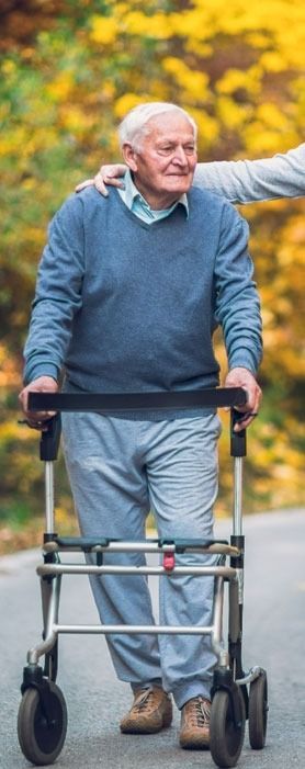 Elderly man smiling while walking with a walker on a path lined with autumn leaves.