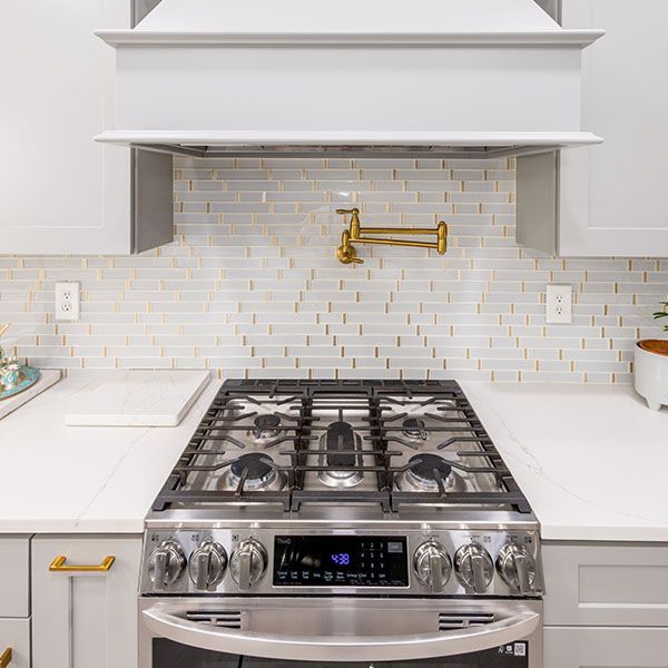 Modern kitchen with stainless steel gas range, white countertops, and brass pot filler under a vent hood