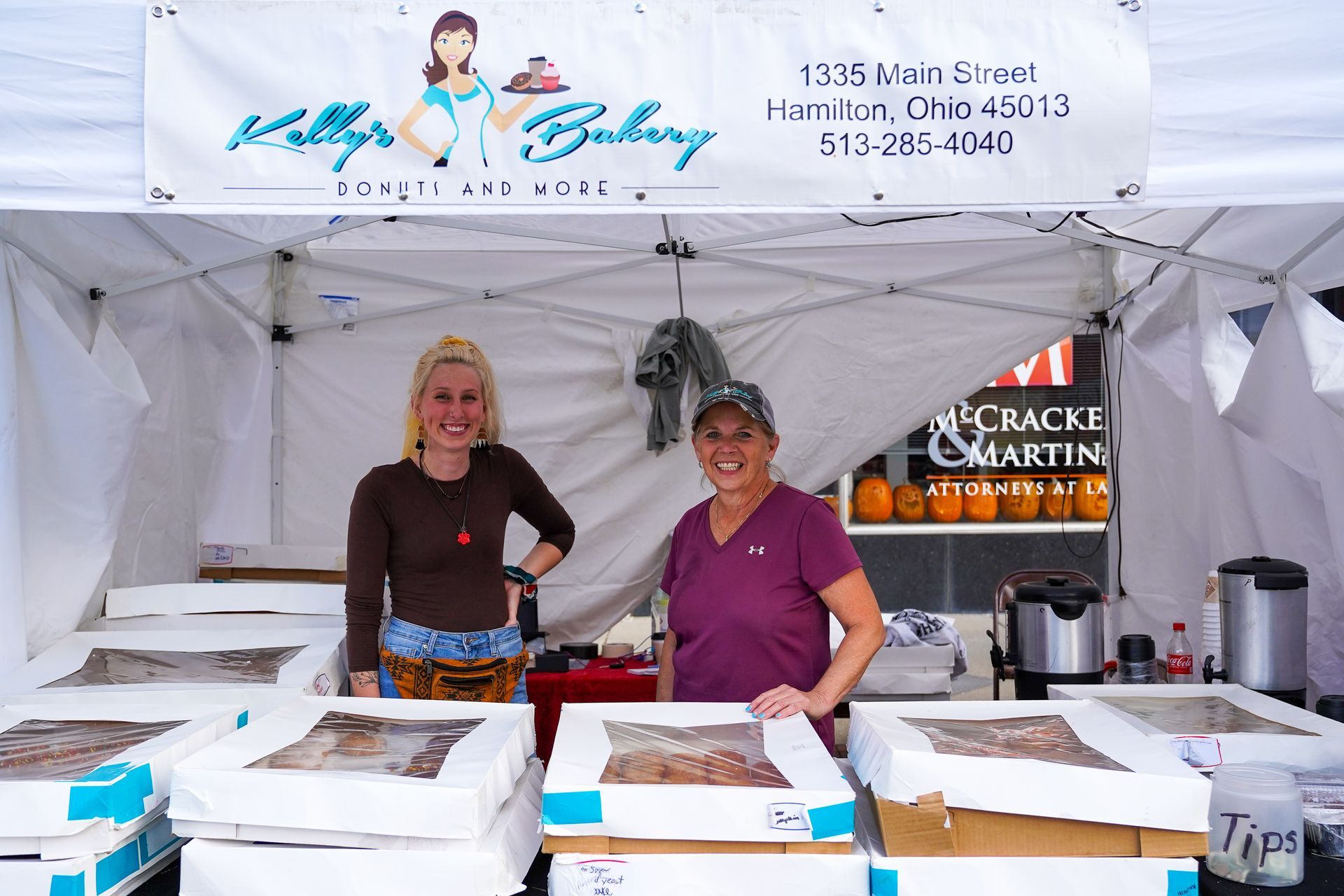 two women standing in front of a kelly 's bakery tent