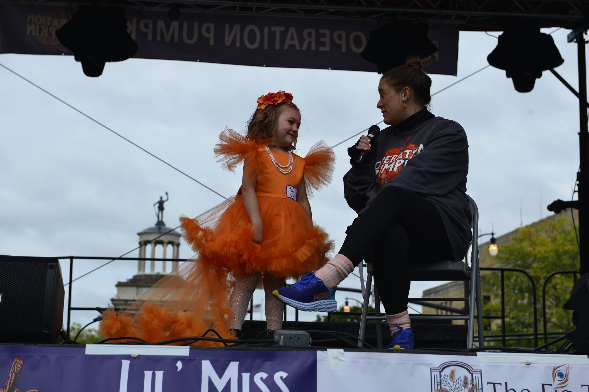 a woman is sitting on a stage talking to a little girl in an orange dress
