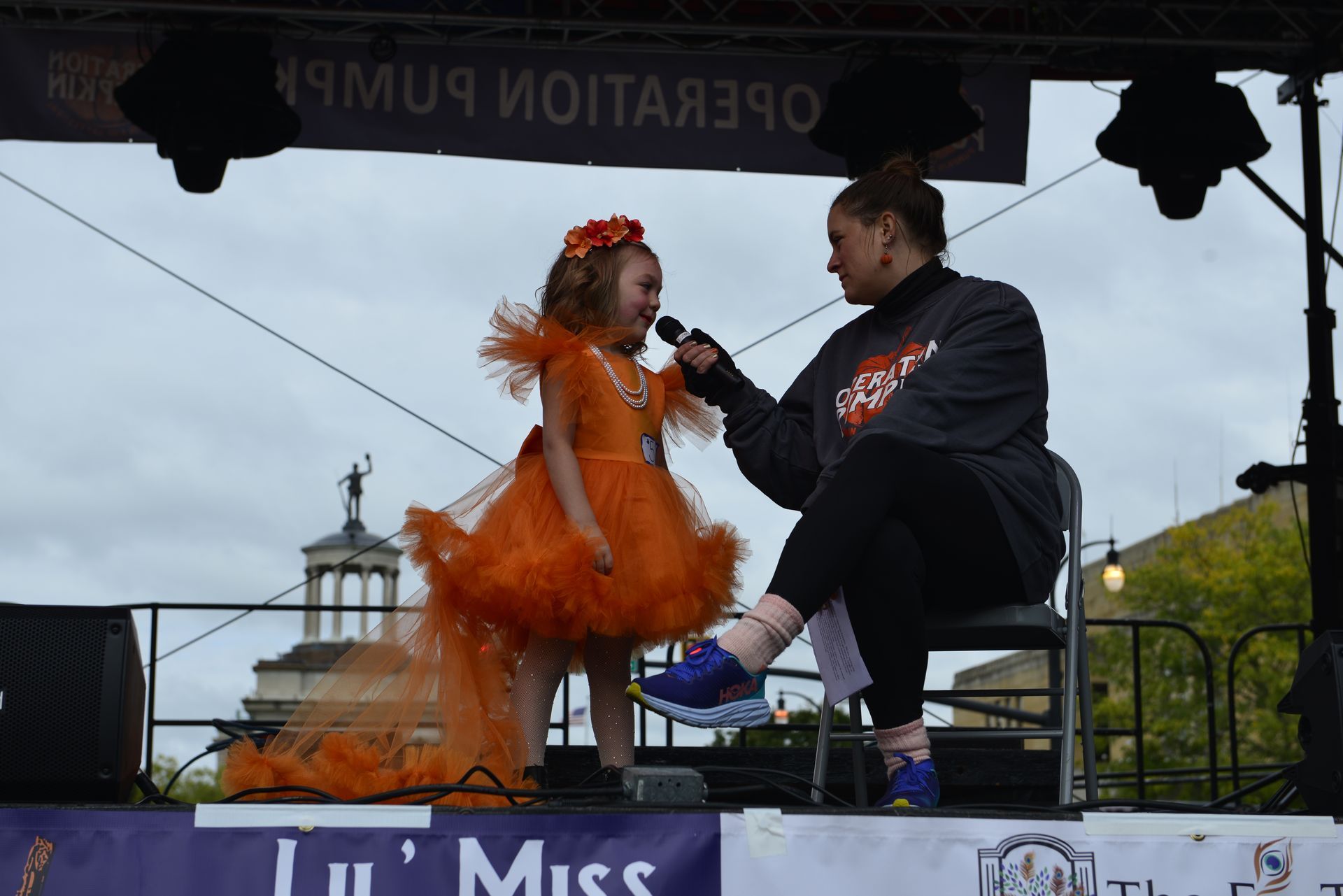 a woman is sitting on a stage talking to a little girl in an orange dress