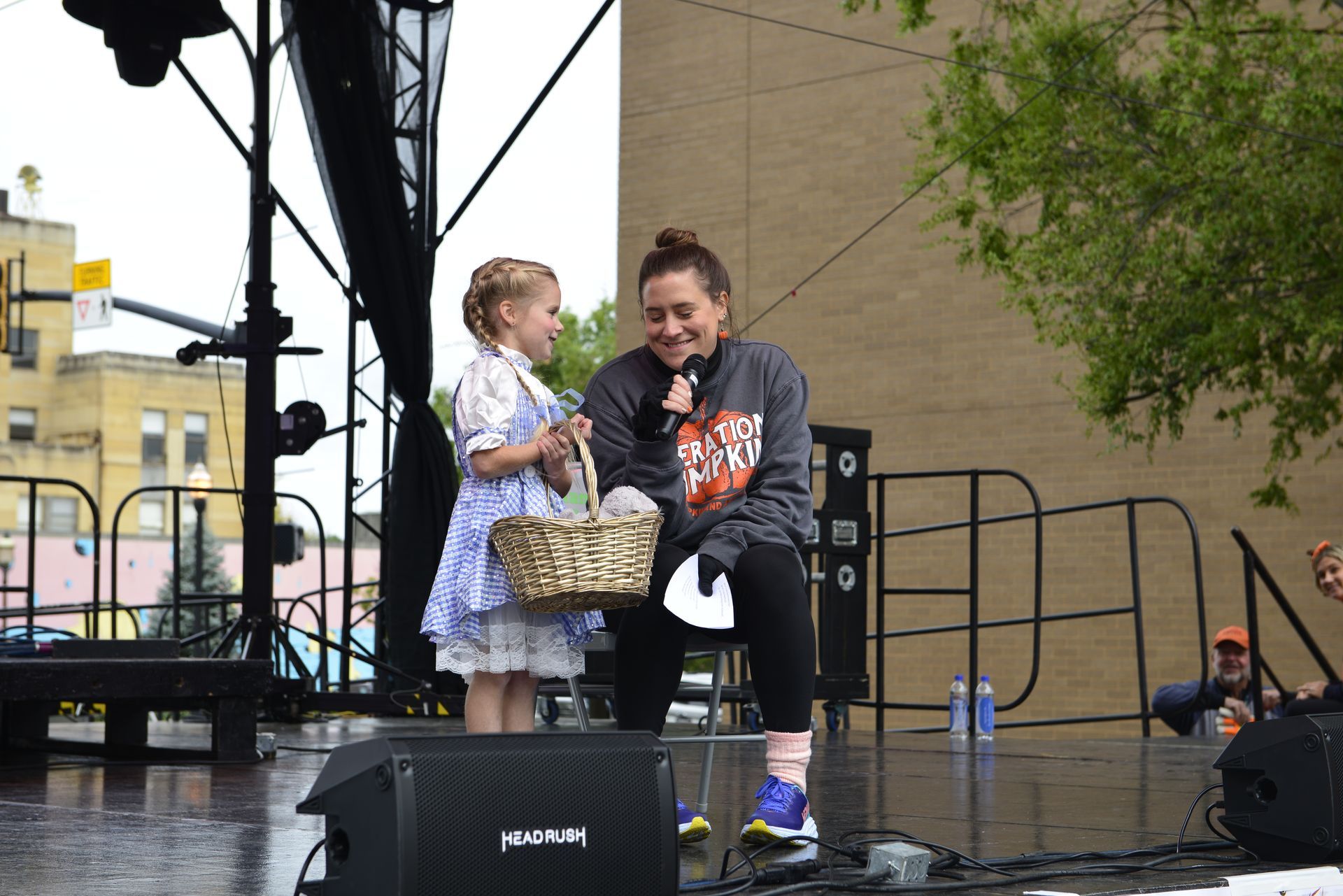 a woman is sitting on a stage talking to a little girl who is holding a basket