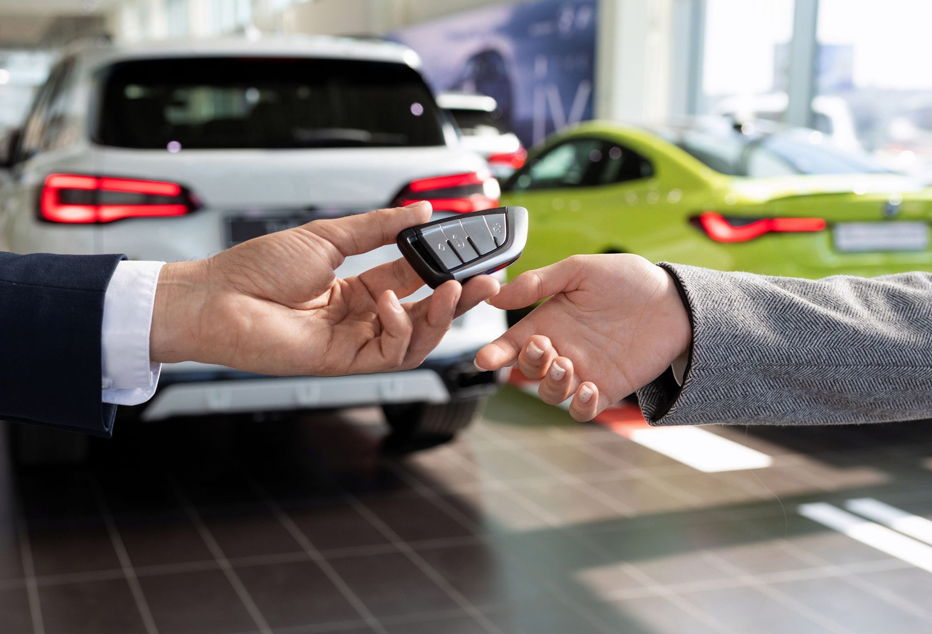 Person handing car keys to another in a car dealership, with two cars in the background.