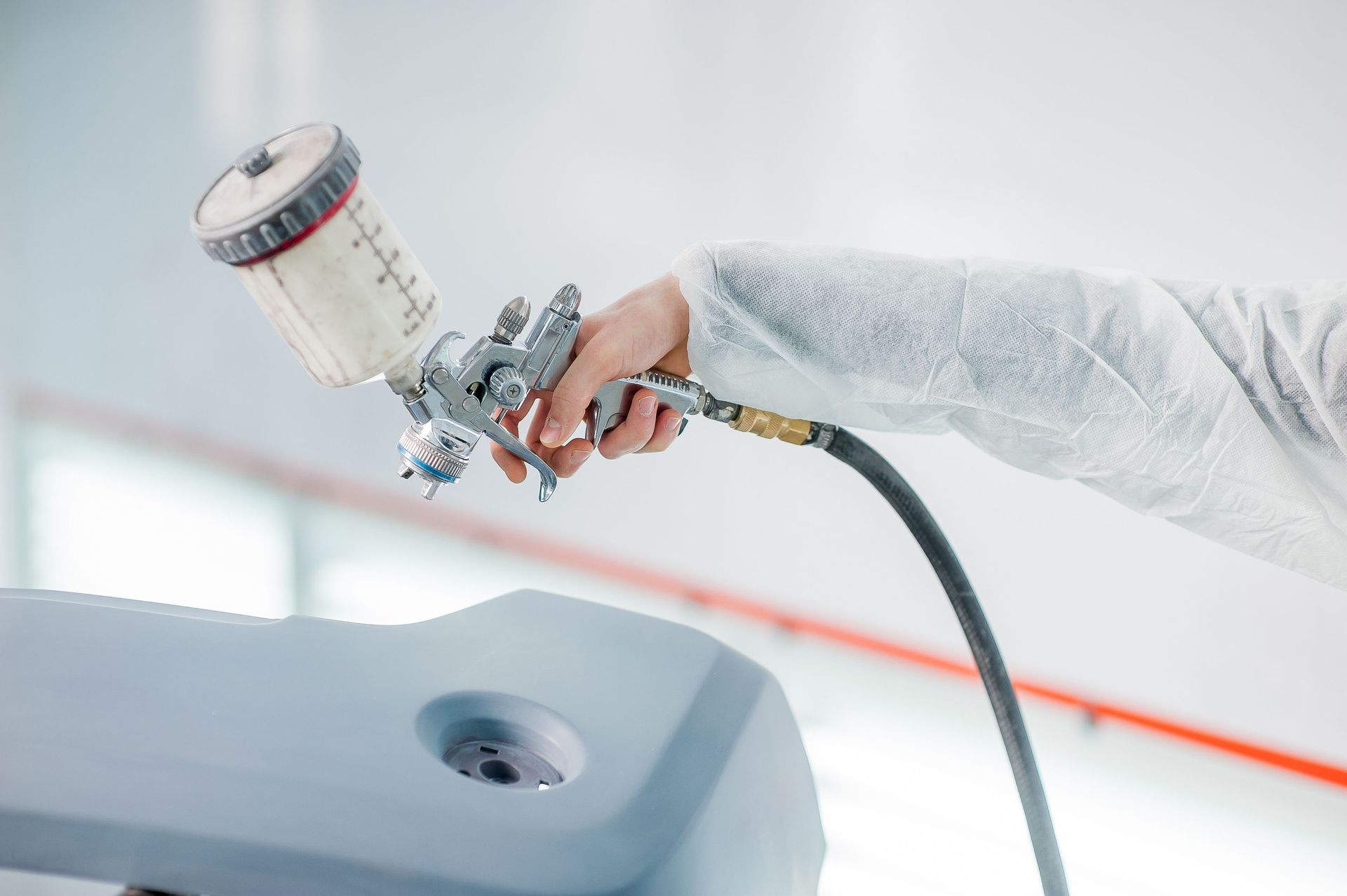 Person in protective suit using a paint sprayer to coat a light gray car part in a workshop.