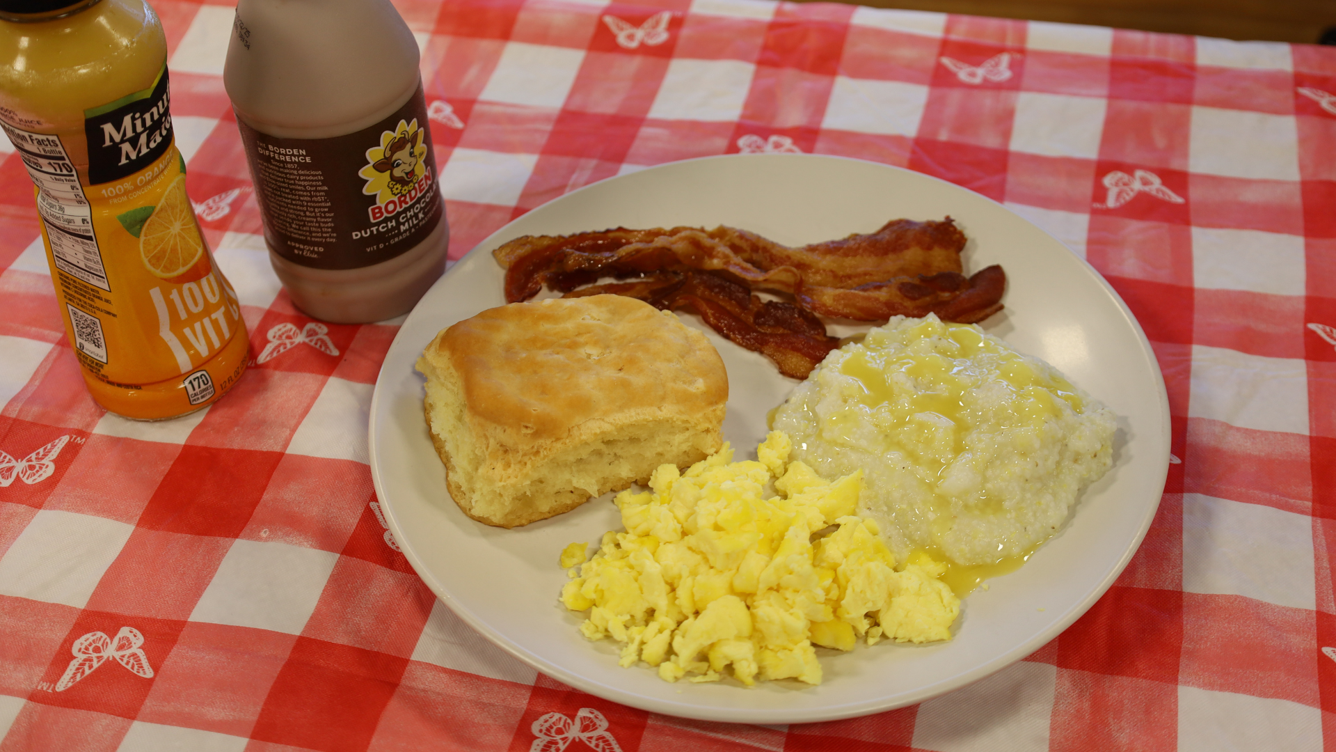 Breakfast plate with biscuit, bacon, scrambled eggs, and grits on a red-checkered tablecloth