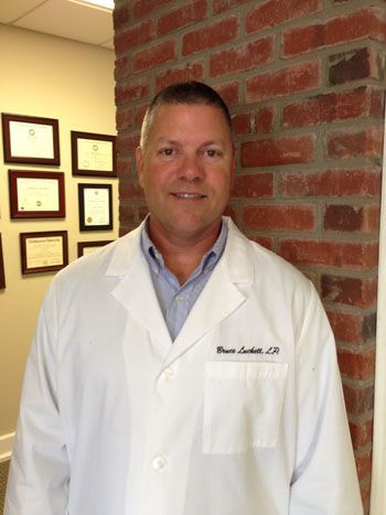 Man in white lab coat smiles, stands by a brick wall and a hallway with diplomas.