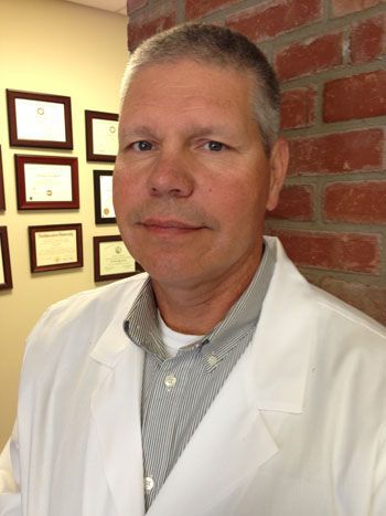 Man in white lab coat smiles, stands by a brick wall and a wall with framed documents.