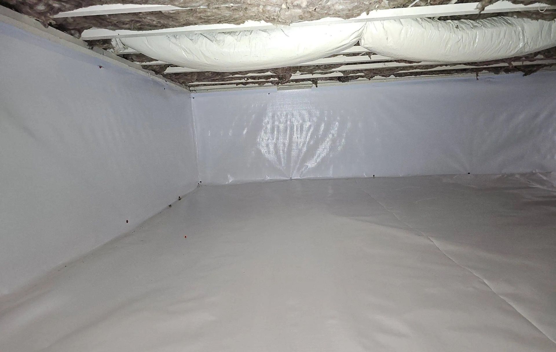 A crawl space interior featuring white vapor barrier lining the walls and floor, with insulation installed overhead.