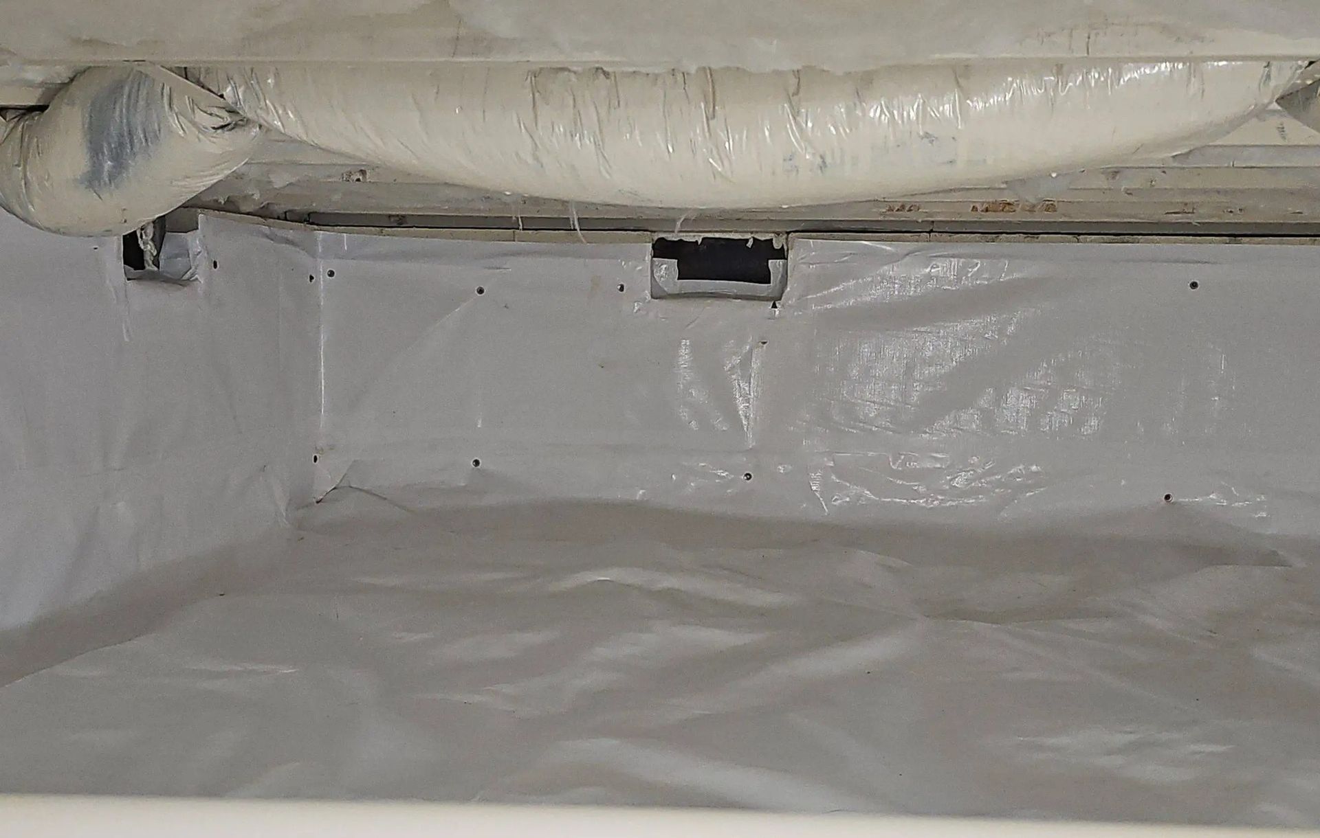 A crawl space featuring white vapor barrier sheeting on the walls and floor, with spray foam insulation on the floor joists.
