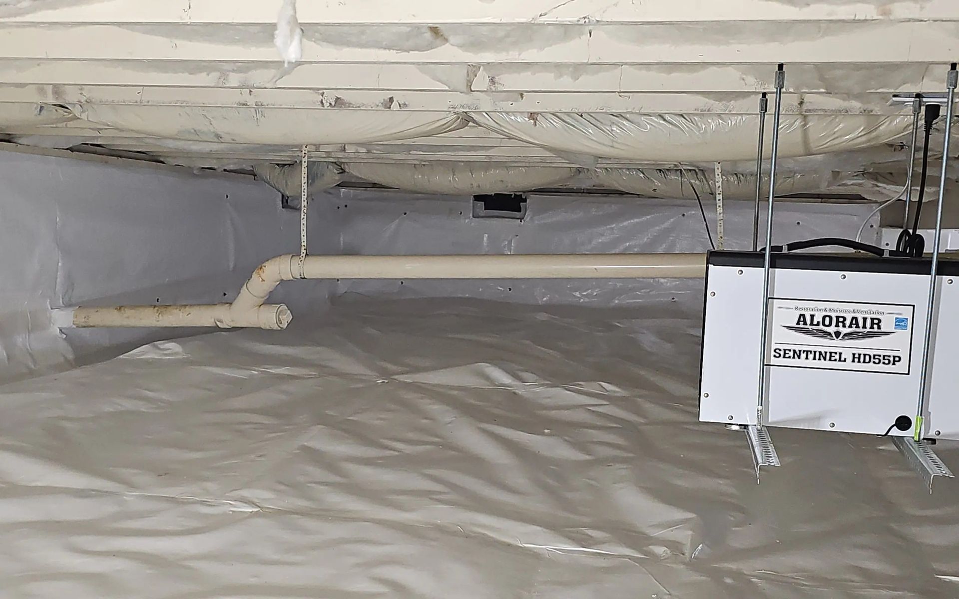 A crawlspace with a white plastic vapor barrier, overhead spray foam insulation, and an AlorAir dehumidifier unit.