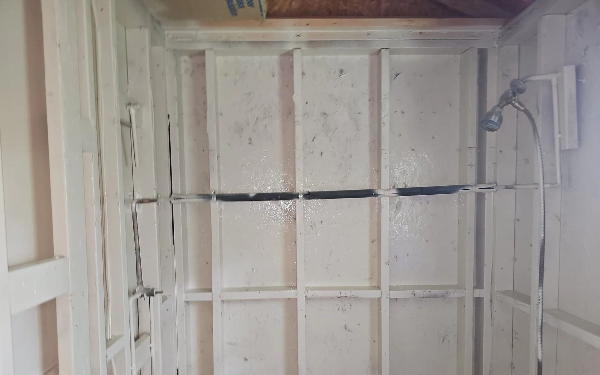 An unfinished white-painted shower stall interior with exposed wooden wall studs and a wall-mounted showerhead.