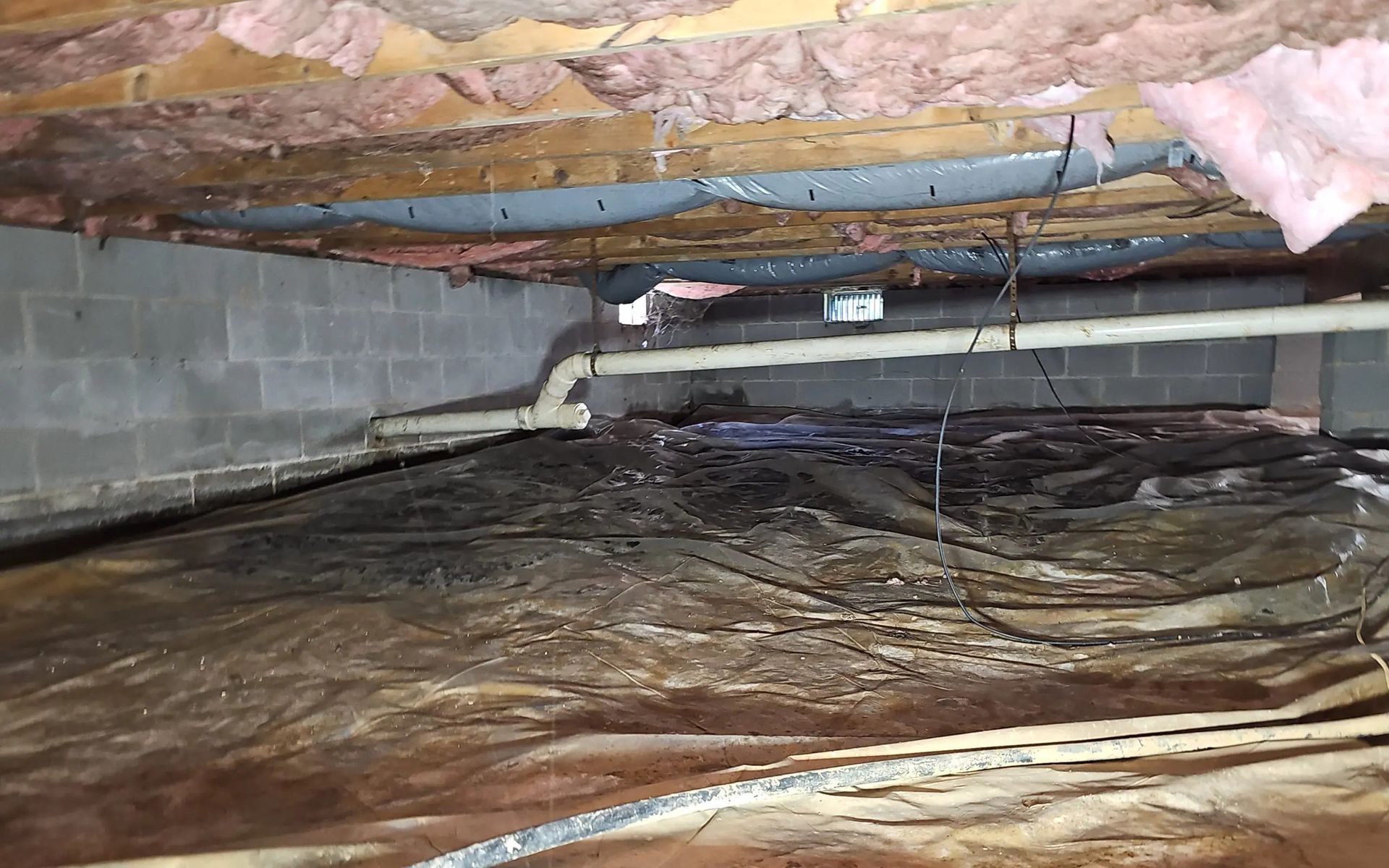 Crawl space with pink insulation between floor joists, a white drain pipe, and a dark, damp ground vapor barrier.