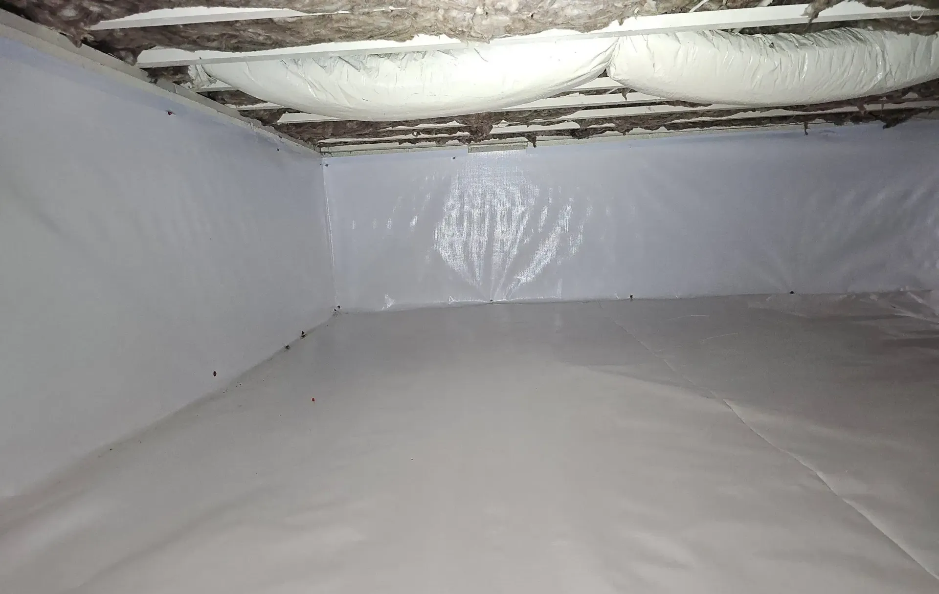 A crawl space encapsulated with white plastic vapor barrier sheeting on the walls and floor, with spray foam insulation.