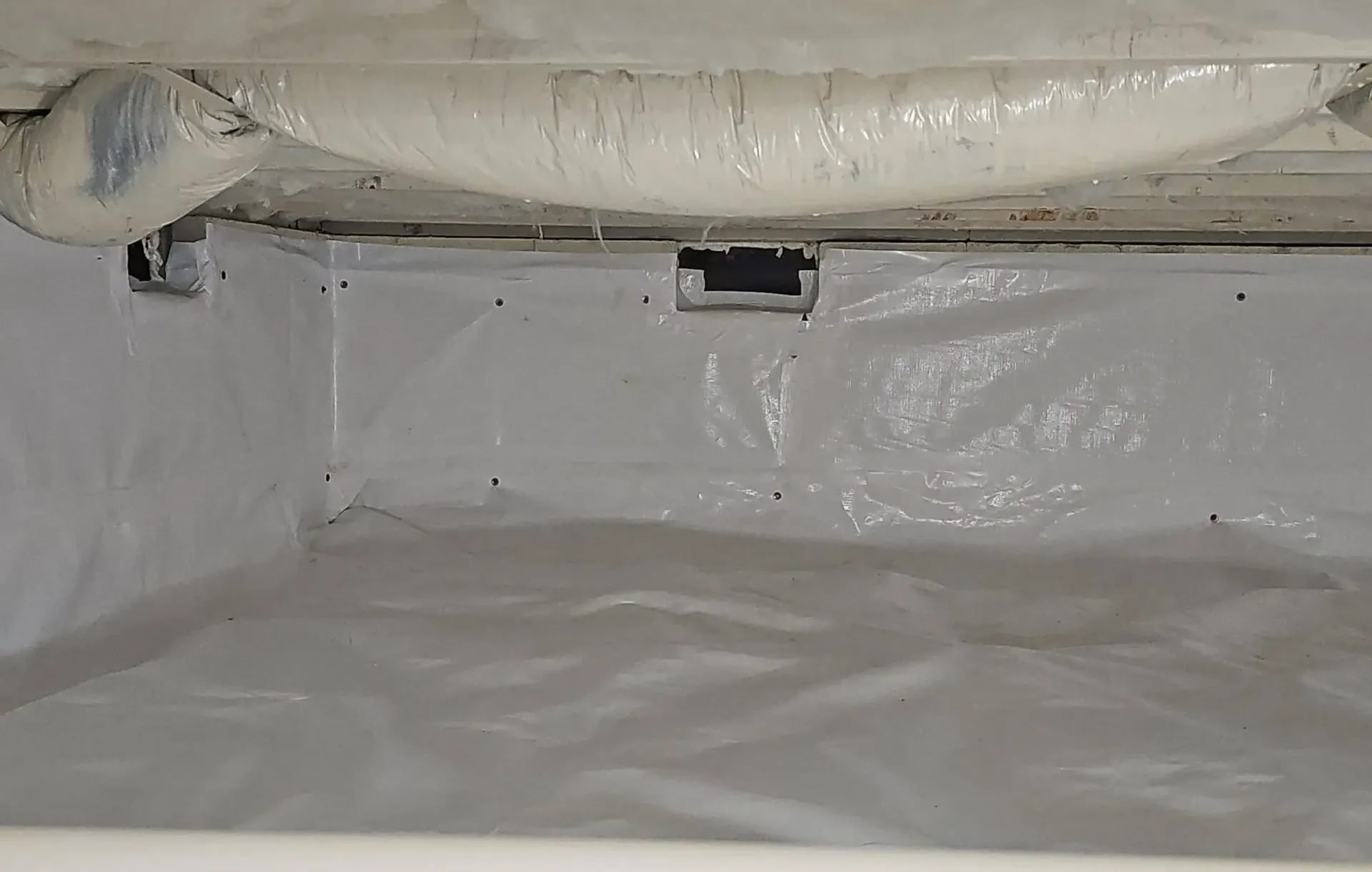 A white vapor barrier installed in a crawl space, covering the ground and walls, with visible vents in the foundation.