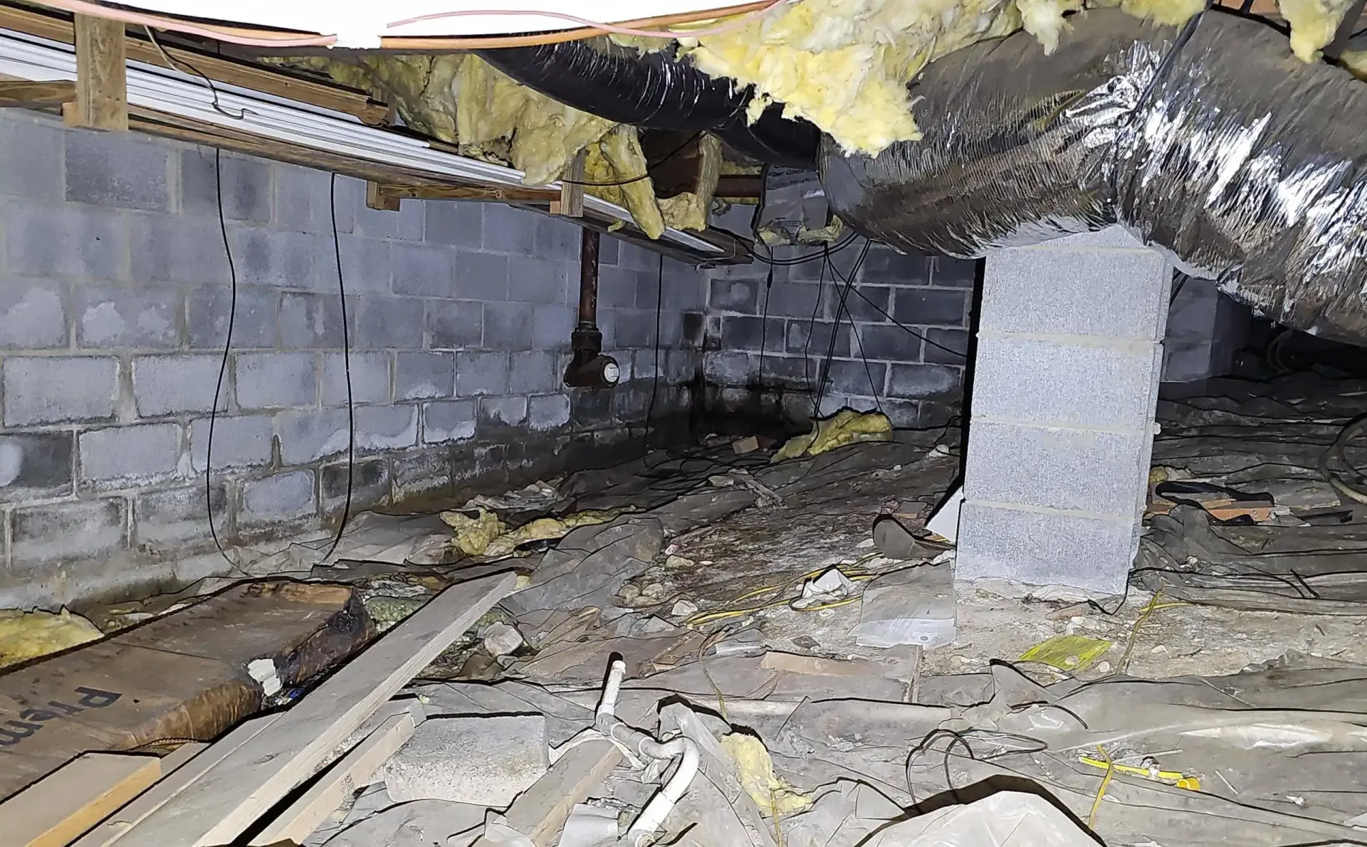 A crawl space with cinder block walls, exposed insulation, a large HVAC duct, and debris covering the ground.