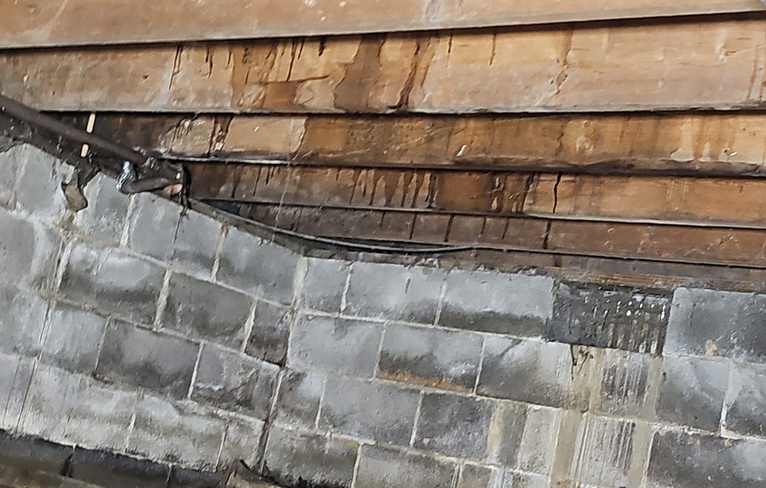 A view looking up at a concrete block wall meeting dark, weathered wooden roof decking showing signs of water damage.