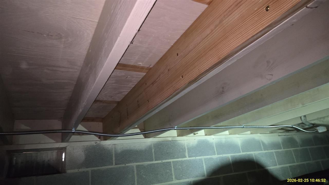Low-angle view of floor joists and electrical wiring running along a cinder block foundation wall in a crawl space.