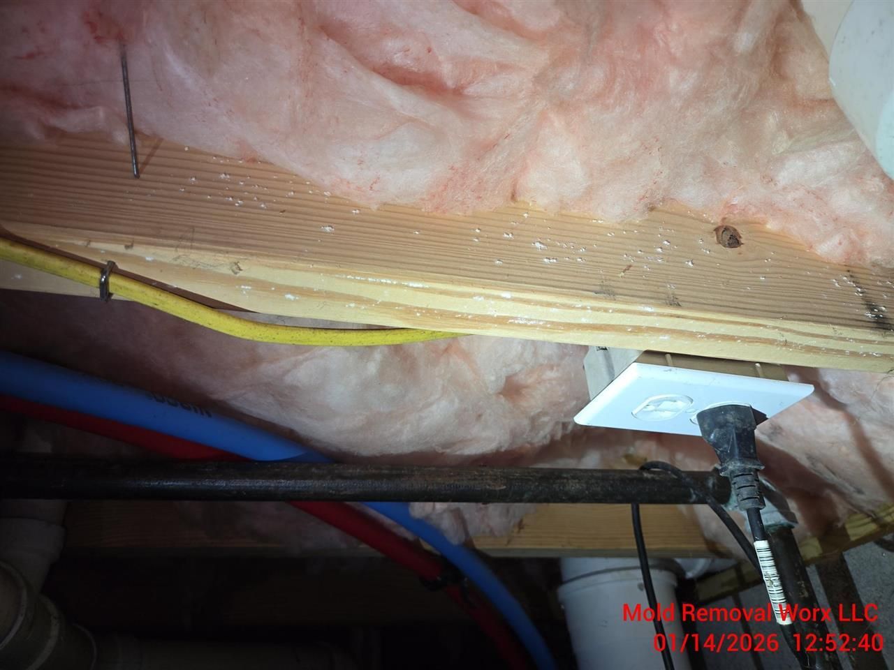 Exposed wooden floor joists and pink fiberglass insulation in a ceiling, with a yellow cable and an electrical outlet.