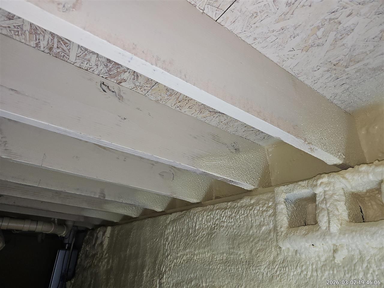 View of wooden floor joists above a foundation wall, both insulated with spray foam in an unfinished basement.