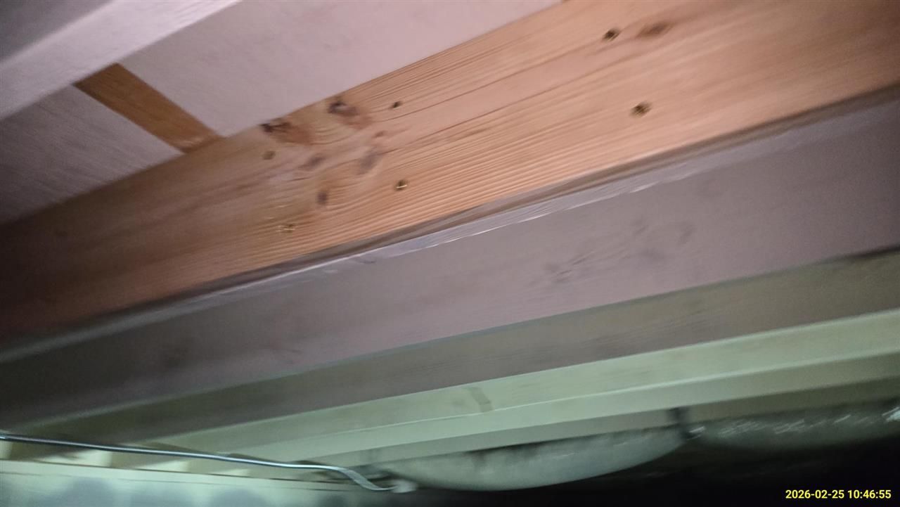 A wooden beam and a grey steel I-beam running horizontally along a ceiling, viewed from below in a crawl space.