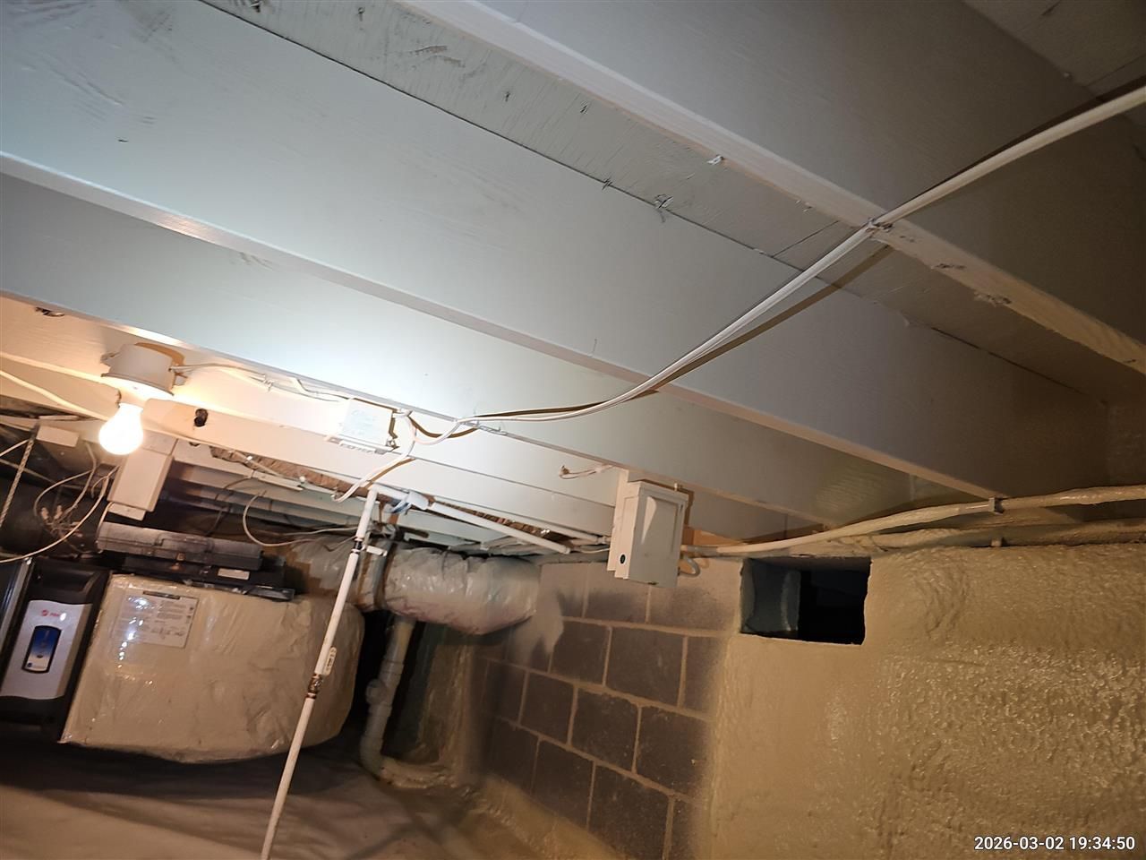 An unfinished basement ceiling with exposed white wooden joists, hanging electrical wiring, and a light bulb.