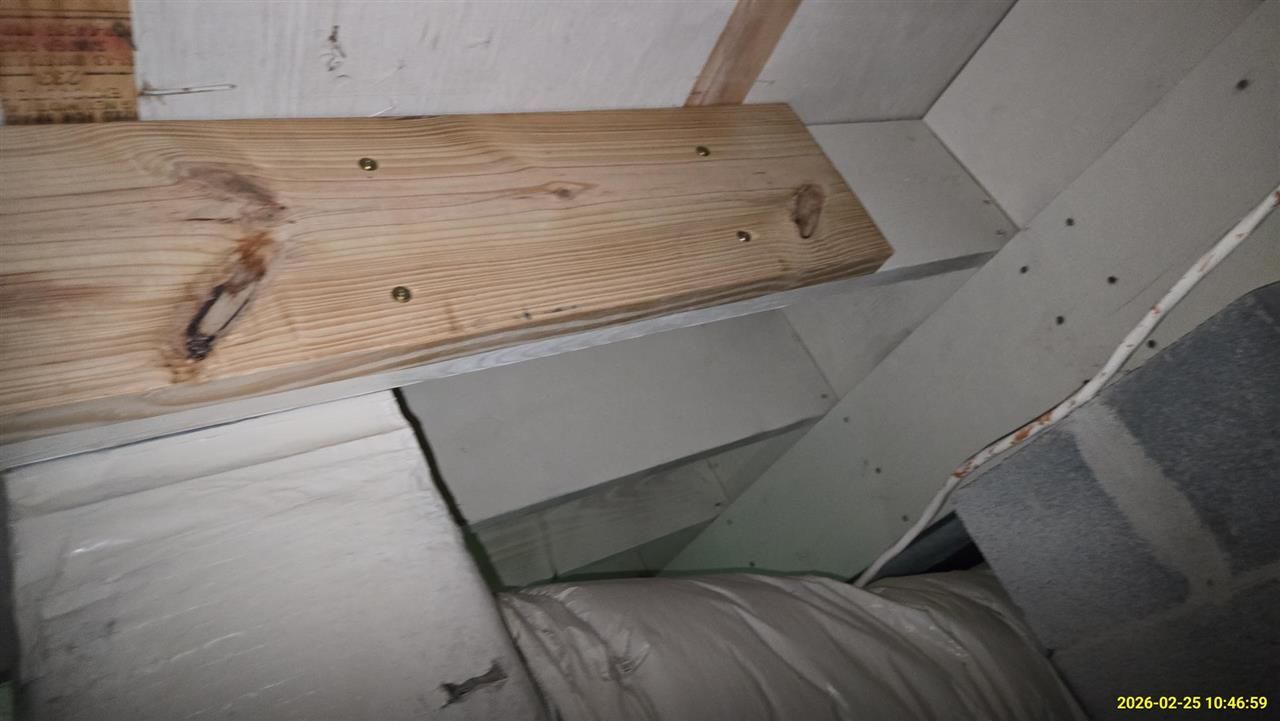 A wooden board is bolted to a painted white joist in an attic, near insulated ducts and structural framing.