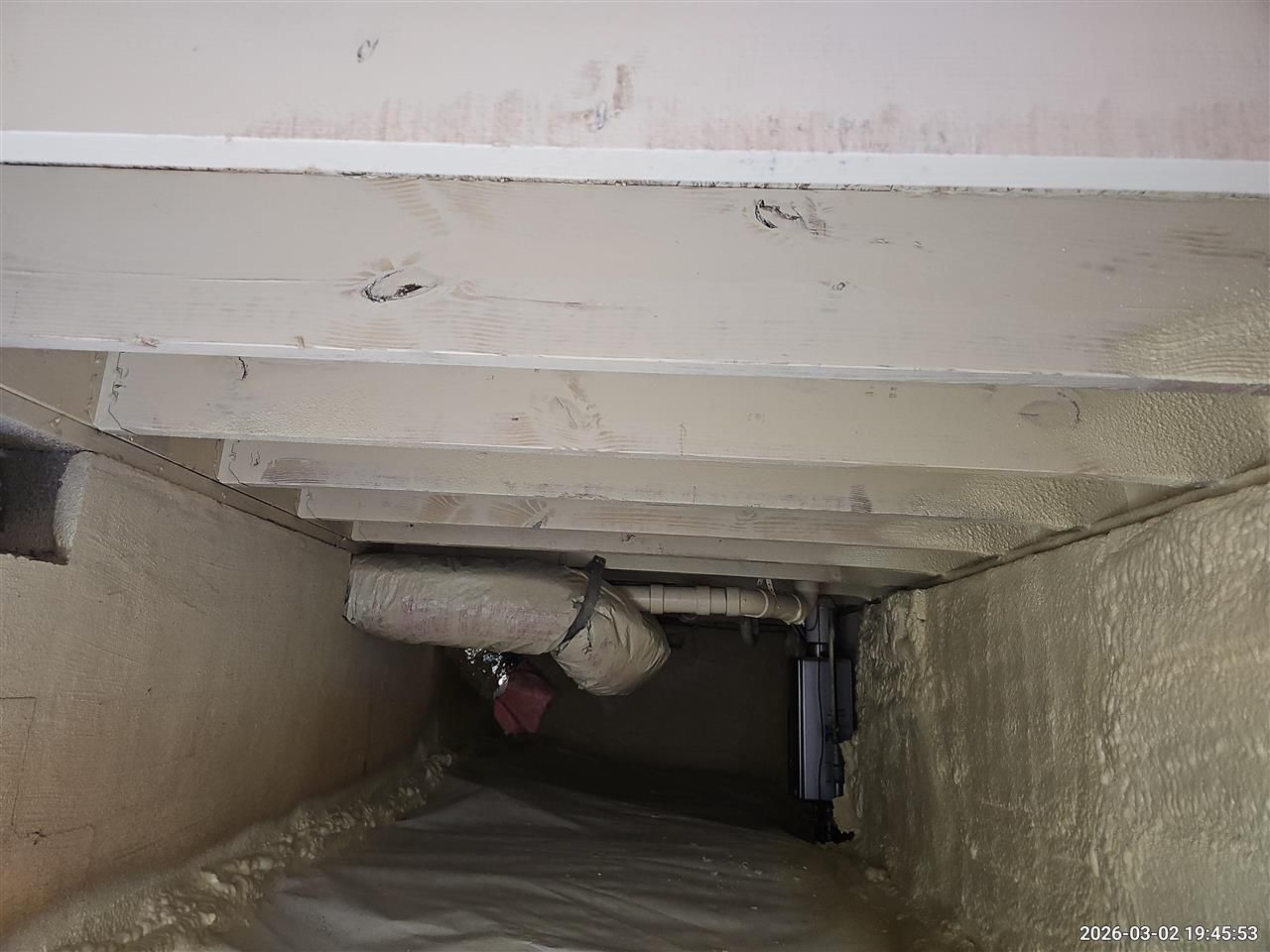 A low-angle view of a crawl space showing wooden floor joists above a vapor barrier with foam-insulated walls.