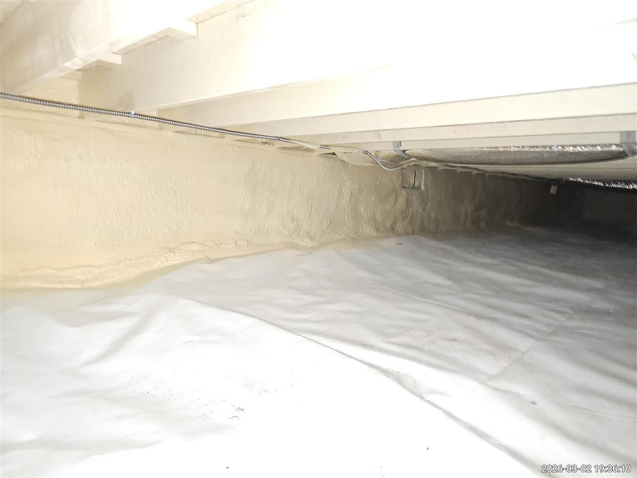 A crawlspace with foam insulation applied to the rim joist and a white vapor barrier covering the ground.