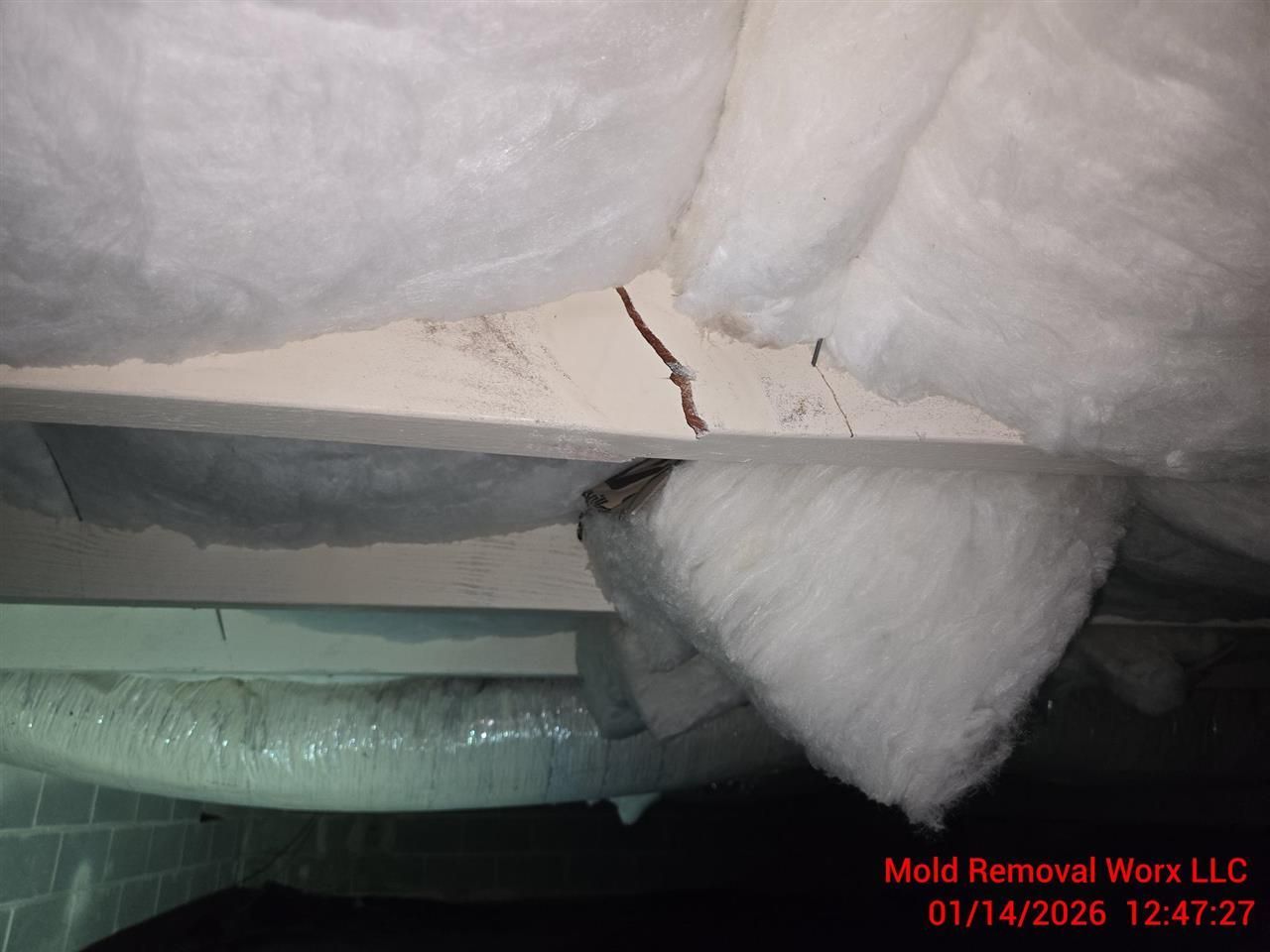 A crawl space showing a structural crack in a wooden ceiling joist surrounded by fiberglass insulation.