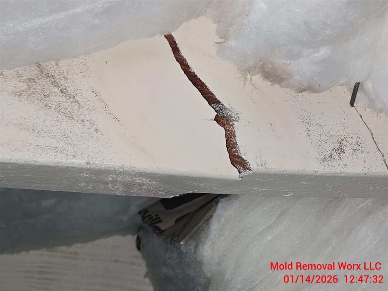 A cracked, light-colored drywall panel with visible brown rust-like staining, surrounded by white fiberglass insulation.