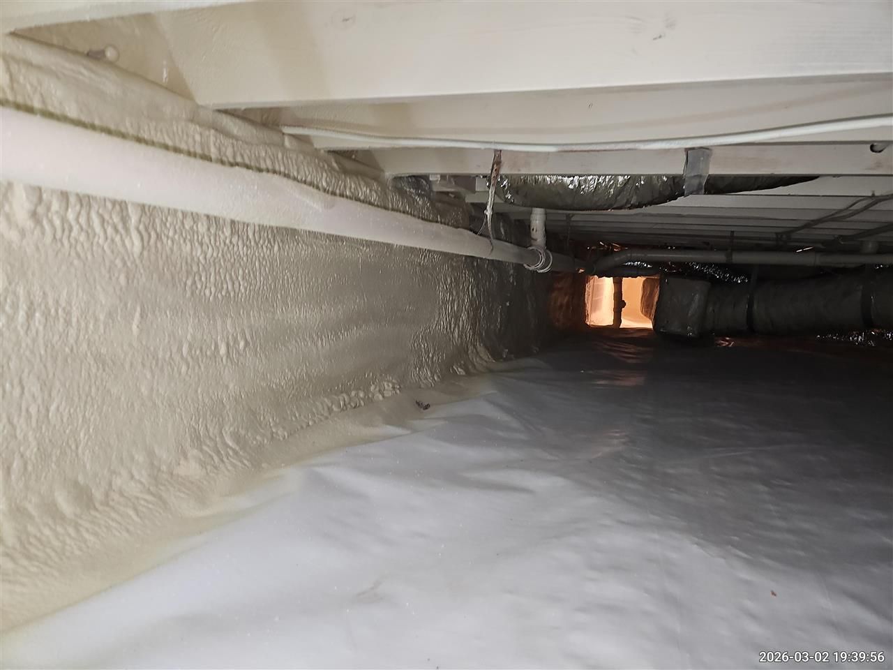 A crawl space with white spray foam insulation on the foundation walls and a white vapor barrier covering the ground.