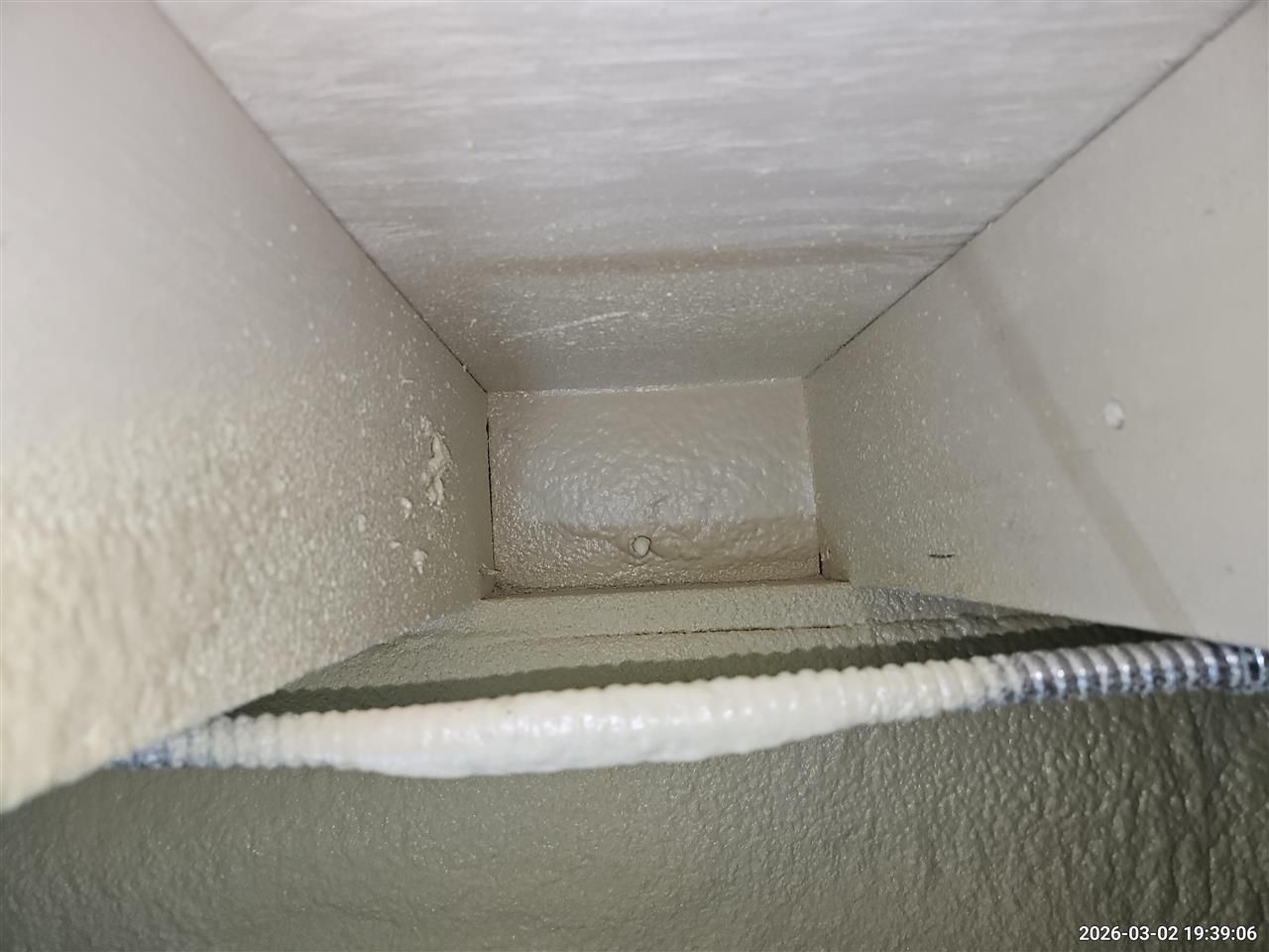 A close-up view of a rectangular ventilation duct, showing textured interior walls and a corrugated metal pipe.